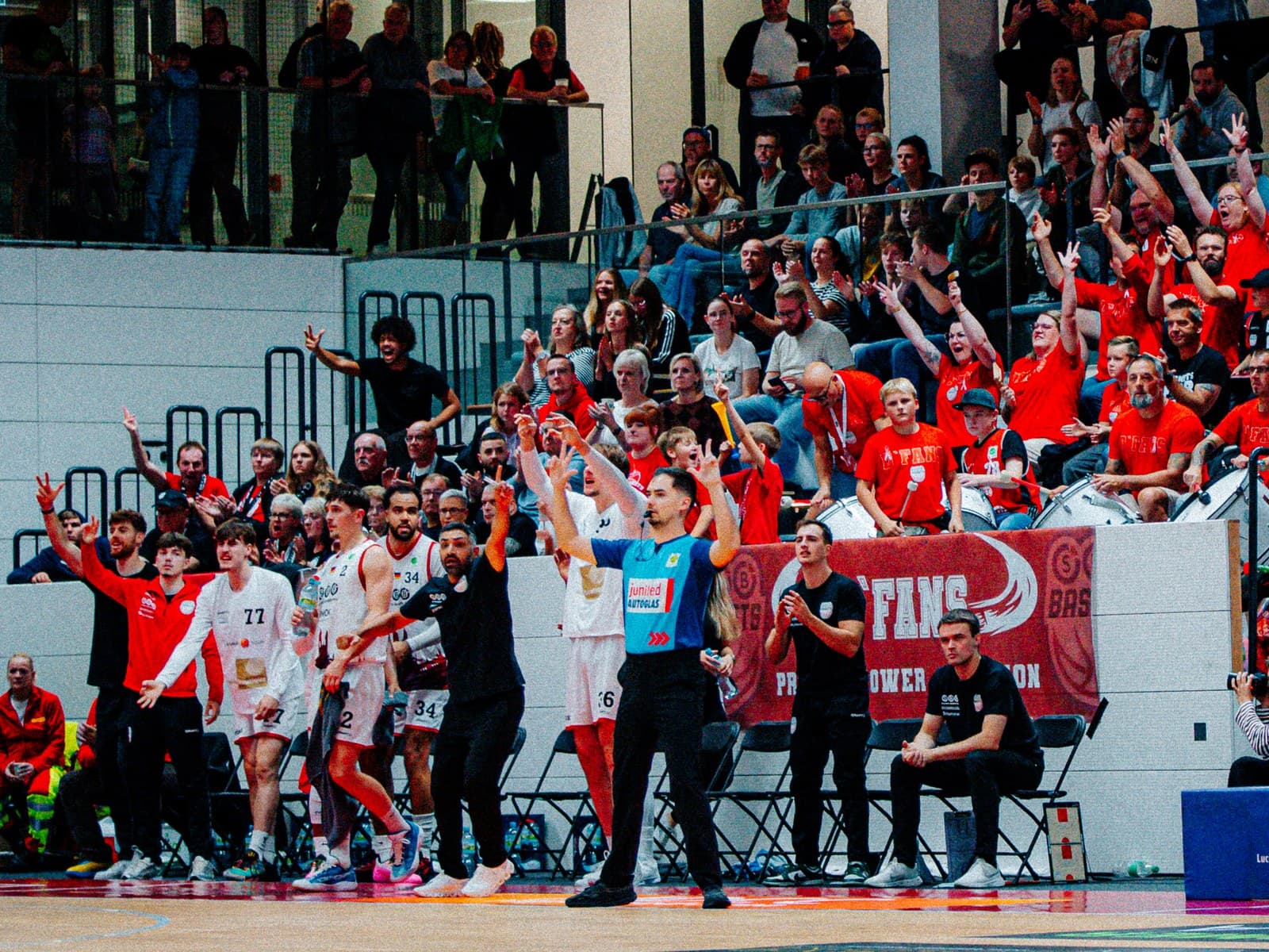 Heimspiel in der Wolfgang-Lakenmacher-Halle: Spieler und Trainerstab der SBB Börde Baskets jubeln an der Seitenlinie, dahinter feiert der Fanblock in Rot.
