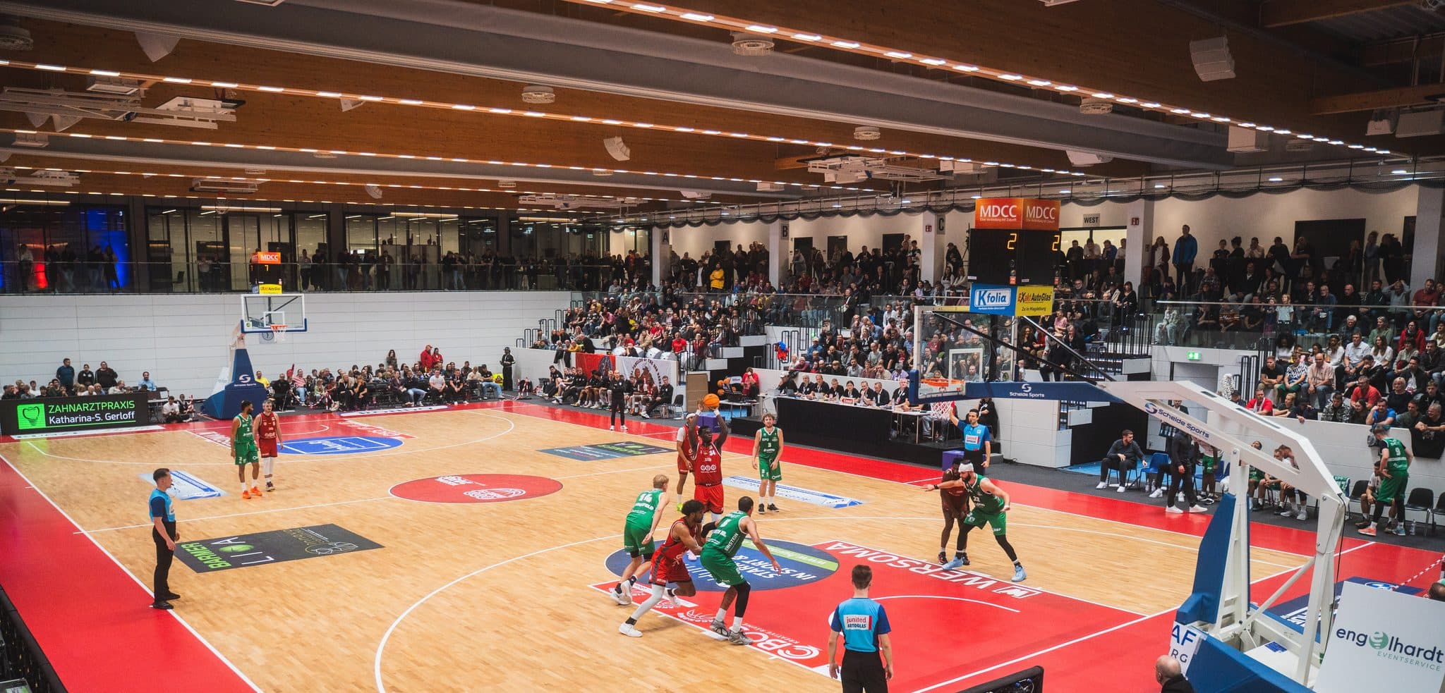 Blick von der Tribüne in die Wolfgang-Lakenmacher-Halle während eines Heimspiels: das rot-helle Spielfeld, die Mannschaften in Aktion, besetzte Ränge.